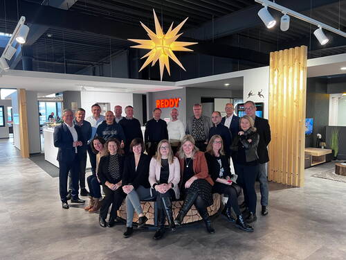 Gruppe von Geschäftsleuten posiert lächelnd in einem modernen Showroom mit großer leuchtender Sternlampe an der Decke und dem roten Schriftzug „REDDY“ im Hintergrund.