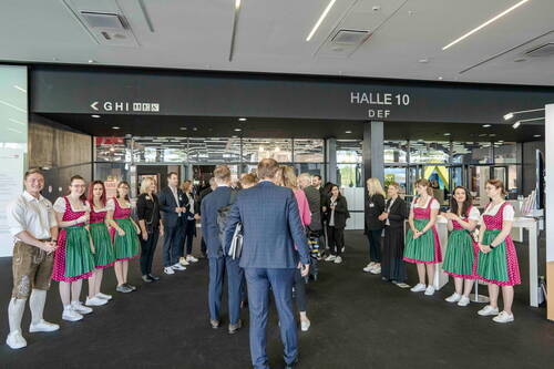 Besuchergruppe in Businesskleidung wird in einer Messehalle von jungen Personen in traditionellen Trachten begrüßt, die links und rechts einen Spalier bilden.