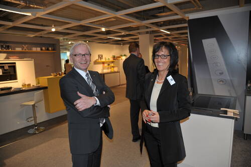 Zwei elegant gekleidete Geschäftsleute unterhalten sich lächelnd in einer modernen Küchenausstellung während einer Veranstaltung.