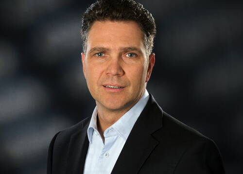Mittelalterlicher Mann im dunklen Anzug und hellblauem Hemd blickt ernst in die Kamera vor unscharf dunklem Hintergrund