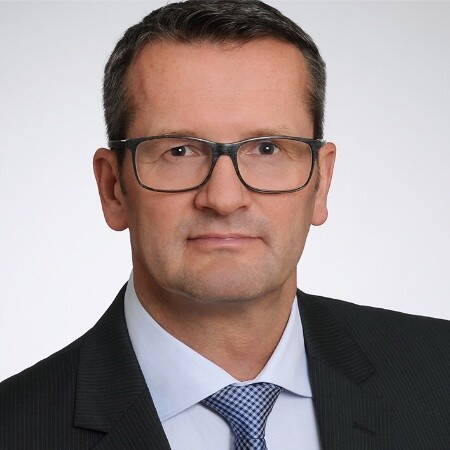 Mittelalterlicher Mann mit kurzem dunklem Haar, Brille und ernstem Gesichtsausdruck im dunklen Anzug mit hellblauem Hemd und gemusterter Krawatte vor neutralem Hintergrund