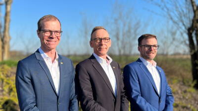 Drei lächelnde Männer in Anzügen mit Brille posieren nebeneinander im Freien vor unscharfer Landschaft bei blauem Himmel