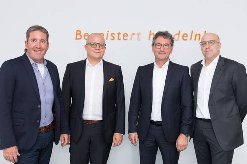 Vier Geschäftsleute in Anzügen stehen nebeneinander vor einer weißen Wand mit orangefarbener Aufschrift und blicken lächelnd in die Kamera.
