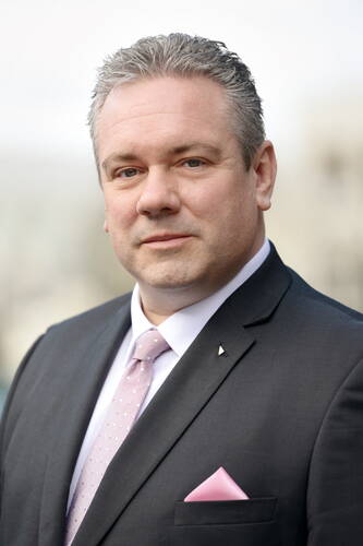 Mittelalterlicher Mann im dunkelgrauen Anzug mit rosa Krawatte und Einstecktuch blickt ernst in die Kamera vor unscharfem, hellem Hintergrund.