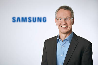 Lächelnder Geschäftsmann mit Brille im Anzug vor einem unscharfen Samsung‑Logo auf hellem Hintergrund