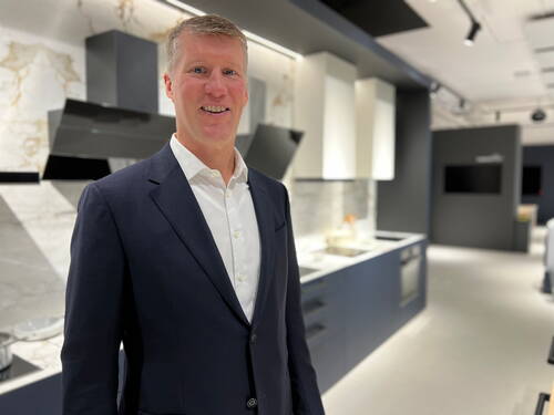 Mittelalter Mann im dunklen Anzug steht lächelnd in einer modernen, hell beleuchteten Ausstellungsküche mit Einbaugeräten und Marmor-Rückwand.