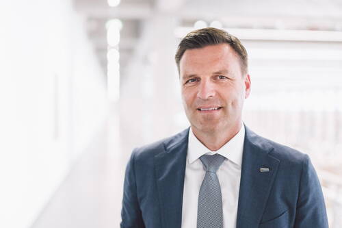 Mittelalterlicher Geschäftsmann im blauen Anzug mit Krawatte steht lächelnd in einem hellen, modernen Büroflur.