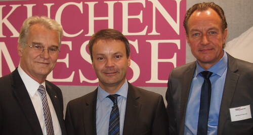 Drei Geschäftsleute im Anzug posieren lächelnd vor einem Messestand mit großem pinkem Schriftzug im Hintergrund