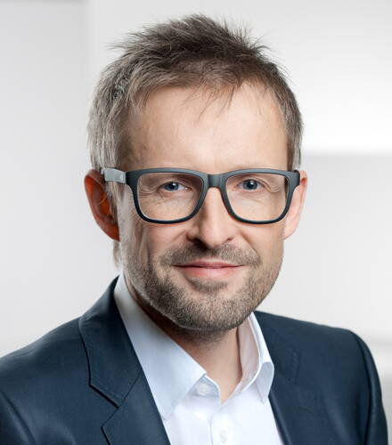 Mittelalterlicher Mann mit kurzem, strubbeligem Haar, dunkler Brille und Dreitagebart lächelt freundlich in die Kamera und trägt ein dunkles Sakko über einem weißen Hemd vor hellgrauem Hintergrund.
