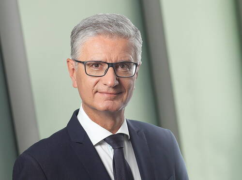 Älterer Mann im Anzug mit Brille und grauen Haaren blickt ruhig und direkt in die Kamera vor unscharfem, modernem Hintergrund.