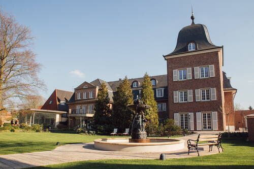 Historische Backsteinvilla mit Türmchen, Springbrunnen und Parkbänken in einem gepflegten Garten bei klarem Himmel