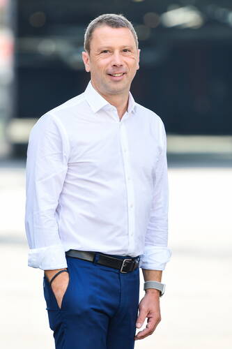 Mittelalterlicher Mann in weißem Hemd und blauer Hose steht lächelnd im Freien mit einer Hand in der Hosentasche.