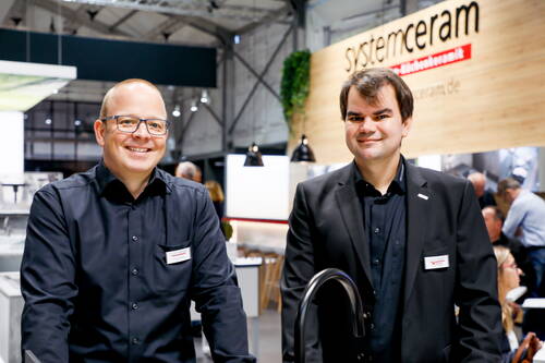 Zwei Mitarbeiter im schwarzen Hemd und Sakko lächeln an einem Messestand von Systemceram vor einem modernen Küchenbereich in einer Halle.