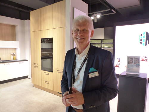 Älterer Mann im Anzug mit Namensschild lächelt in einer modernen Küchenausstellung mit hellen Holzmöbeln und Einbaugeräten.
