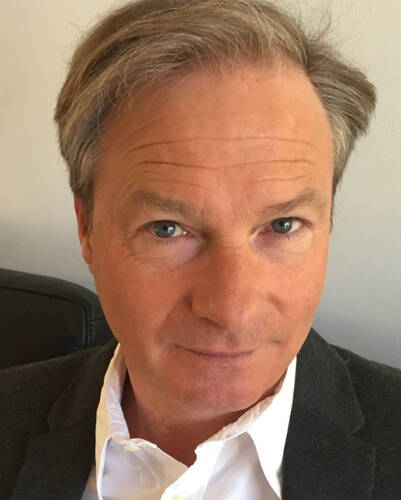 Mittelälterlicher Mann mit grauem Haar und ernstem Blick sitzt in einem dunklen Sakko und weißem Hemd vor einer hellen Wand und schaut direkt in die Kamera.