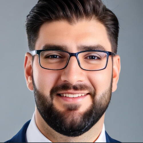 Junger Mann mit dunklem Vollbart, Brille und Anzug lächelt freundlich vor neutral grauem Hintergrund.