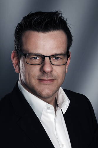 Mittelalterlicher Mann mit dunkler Kurzhaarfrisur und Brille im Business-Outfit mit weißem Hemd und schwarzem Sakko vor neutral grauem Hintergrund