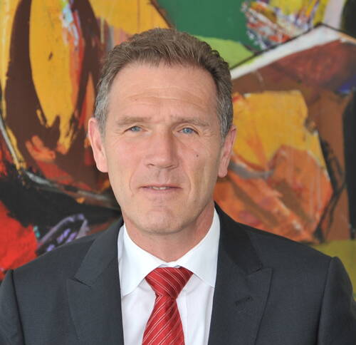 Mittelalterlicher Mann im Anzug mit roter Krawatte blickt ernst in die Kamera, im Hintergrund ein farbenfrohes abstraktes Gemälde