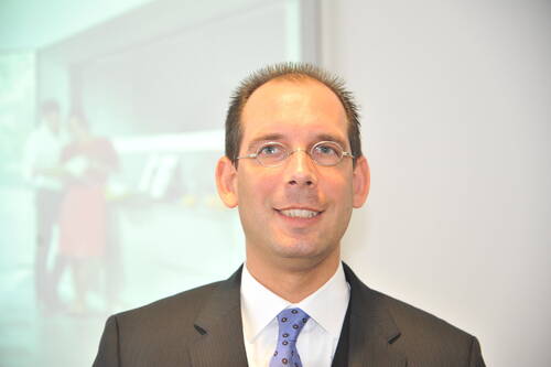 Geschäftsmann mit Brille und Anzug lächelt vor unscharfem Präsentationshintergrund