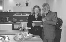 Zwei elegant gekleidete Personen stehen in einer modernen Küche nebeneinander und betrachten gemeinsam ein Tablet über einer Arbeitsfläche mit Obstschalen.