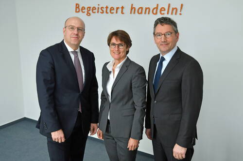 Drei Geschäftsleute in Anzügen stehen lächelnd nebeneinander in einem modernen Büro vor der Wandaufschrift „Begeistert handeln!“.
