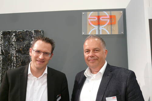 Zwei lächelnde Männer im Business-Outfit posieren vor einer grauen Wand mit dem Firmenlogo eines Küchenherstellers.