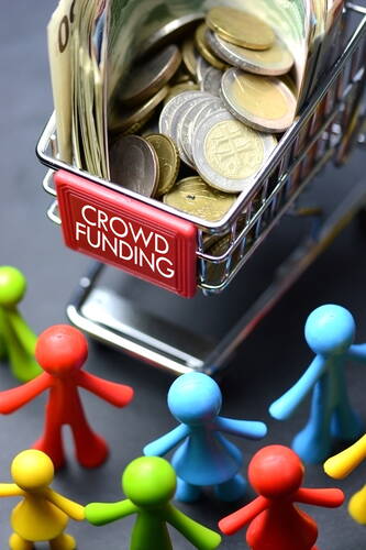 Miniatur-Einkaufswagen mit der Aufschrift „Crowd Funding“ ist mit Scheinen und Münzen gefüllt und wird von bunten, im Kreis stehenden Spielfiguren umringt
