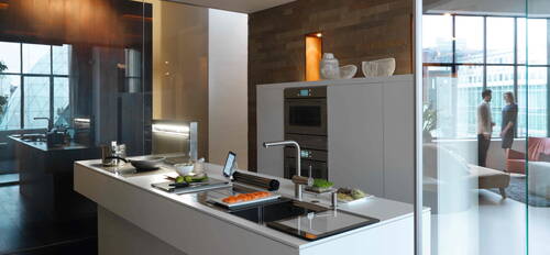 Offene, moderne Designküche mit Kochinsel, frischem Lachs und Gemüse auf der Arbeitsfläche, integriertem Induktionskochfeld und Spülbecken, während im Hintergrund zwei Personen vor bodentiefen Fenstern im Wohnbereich stehen.