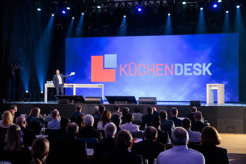 Moderator präsentiert auf großer Bühne vor Publikum eine KüchenDesk-Marke mit leuchtendem Logo auf riesiger LED-Leinwand während einer Konferenz oder Produktvorstellung