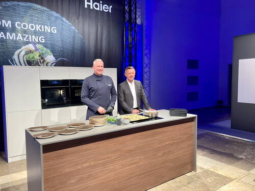 Zwei Männer, ein Koch in dunkler Uniform und ein Manager im Anzug, stehen an einer modernen Kochinsel vor eingebauten Haier-Backöfen und bereiten ein Gericht für eine Bühnenshow mit blau beleuchtetem Hintergrund vor.