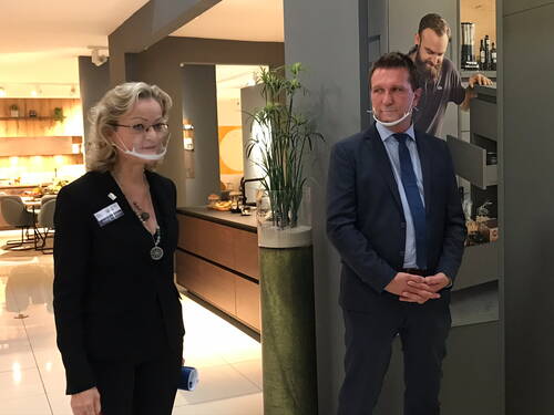 Zwei Geschäftsleute mit transparenten Kinnmasken stehen in einer modernen Ausstellungsküche und hören aufmerksam zu