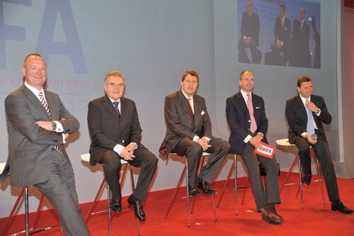Fünf Männer in Anzügen sitzen auf Barhockern auf einer roten Bühne und nehmen an einer Podiumsdiskussion auf einer Messe teil, während hinter ihnen eine Leinwand mit ihrem Live‑Bild projiziert wird.