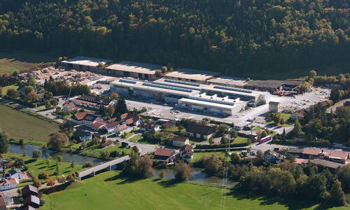 Luftaufnahme eines großen Industrie- und Werksgeländes mit umliegenden Wohnhäusern, Feldern, Fluss und angrenzendem Wald im Hintergrund