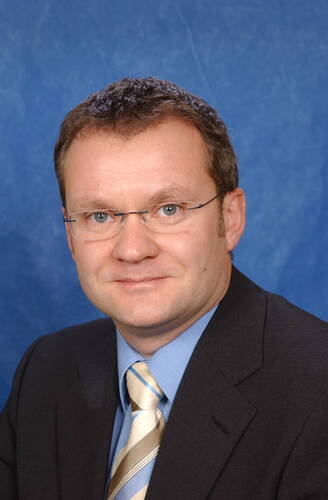 Mittelälterer Mann mit Brille im dunklen Anzug, hellblauem Hemd und gestreifter Krawatte vor blauem Studiohintergrund