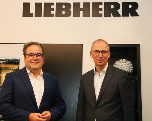 Zwei lächelnde Männer in Anzügen stehen in einem modernen Büro vor einem Regal, über ihnen prangt groß das Firmenlogo „LIEBHERR“.
