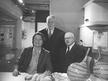 Drei ältere Männer in Anzug und Sakko posieren in einem Ausstellungsraum hinter einem Tisch mit dekorativen Keramikvasen und Stoffobjekten