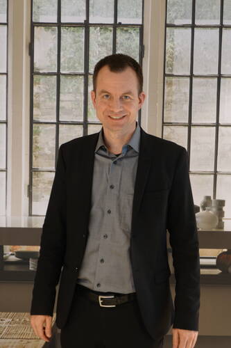 Mittelalter Mann im dunklen Anzug steht lächelnd in einem hellen Raum vor großen Sprossenfenstern.