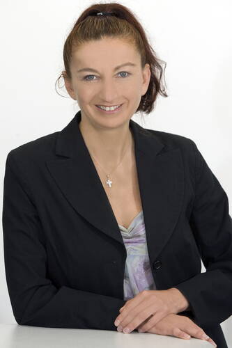 Lächelnde Frau mit Pferdeschwanz in schwarzem Blazer und Kreuzkette sitzt an einem Tisch vor weißem Hintergrund