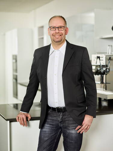 Lächelnder Mann mit Brille im dunklen Sakko und Jeans steht lässig an einer modernen weißen Kücheninsel