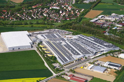 Großes Industrie- und Logistikgelände mit Hallen und Parkplätzen am Rand einer Wohnsiedlung, umgeben von Feldern und Grünflächen aus der Luftperspektive