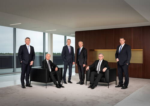 Sechs ältere Männer in dunklen Anzügen posieren als Managementteam in einem modernen, großzügigen Konferenzraum mit Panoramafenstern und Holzvertäfelung, zwei von ihnen sitzen in schwarzen Ledersesseln, vier stehen dahinter.