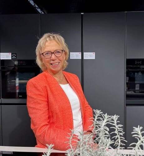 Ältere Frau mit blondem Haar und Brille lächelt in moderner Küche vor dunklen Einbauschränken und trägt ein leuchtend oranges Jackett über weißem Oberteil