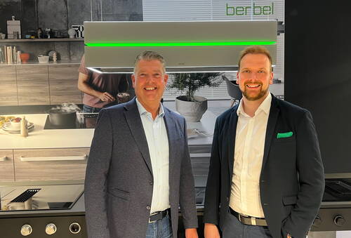 Zwei lächelnde Männer in Anzugjacken stehen in einer modernen Ausstellungsküche vor einer beleuchteten Dunstabzugshaube mit grünem LED-Streifen und dem Logo „berbel“ im Hintergrund.
