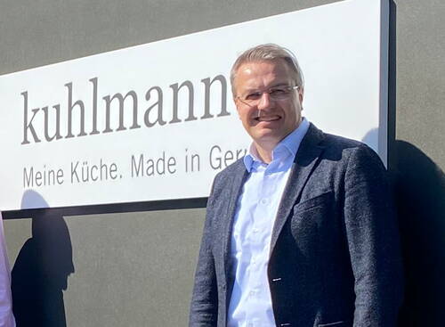 Mittelalter Mann im Sakko lächelt in der Sonne vor einem Firmenschild mit der Aufschrift „kuhlmann – Meine Küche. Made in Germany“
