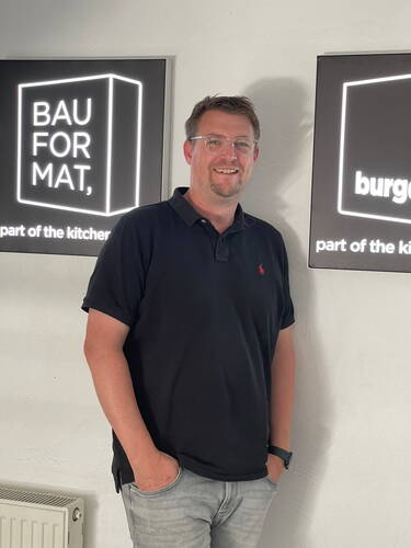 Mann mit Brille und schwarzem Poloshirt steht lächelnd zwischen zwei leuchtenden BAUFORMAT-Wandschildern in einem hellen Raum
