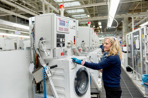 Mitarbeiterin prüft in einer hellen Fabrikhalle eine Waschmaschine an einem automatisierten Teststand mit Kontrollpanelen und Warnleuchten