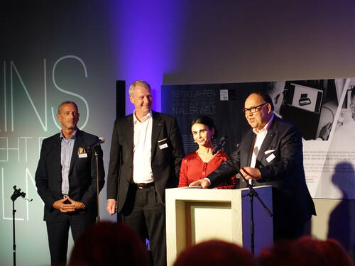 Vier elegant gekleidete Personen stehen auf einer Bühne an Rednerpulten, während ein Mann im Anzug mit Brille in ein Mikrofon spricht und das Publikum bei einer festlichen Veranstaltung adressiert.
