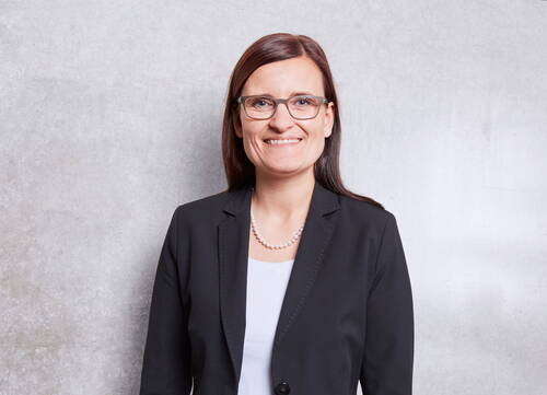 Lächelnde Frau mit Brille in schwarzem Blazer und Perlenkette steht vor einer hellgrauen Betonwand