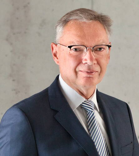 Älterer Geschäftsmann mit Brille im dunkelblauen Anzug und hellblau-weiß gestreifter Krawatte vor neutralem Hintergrund, der ruhig in die Kamera blickt.