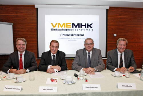 Vier Anzugträger sitzen lächelnd an einem Konferenztisch vor einer Leinwand mit der Aufschrift „VMEMHK Einkaufsgesellschaft mbH – Pressekonferenz“ in einem Tagungsraum.